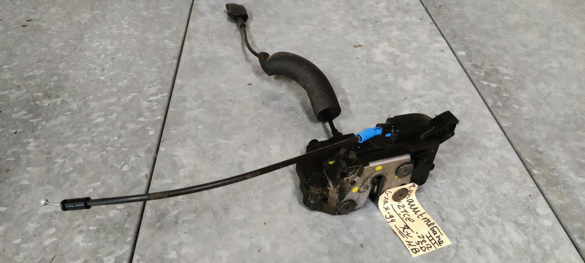 Een zwart autodeurslot met een aangesloten kabel en elektrische connector op een grijze vloer.