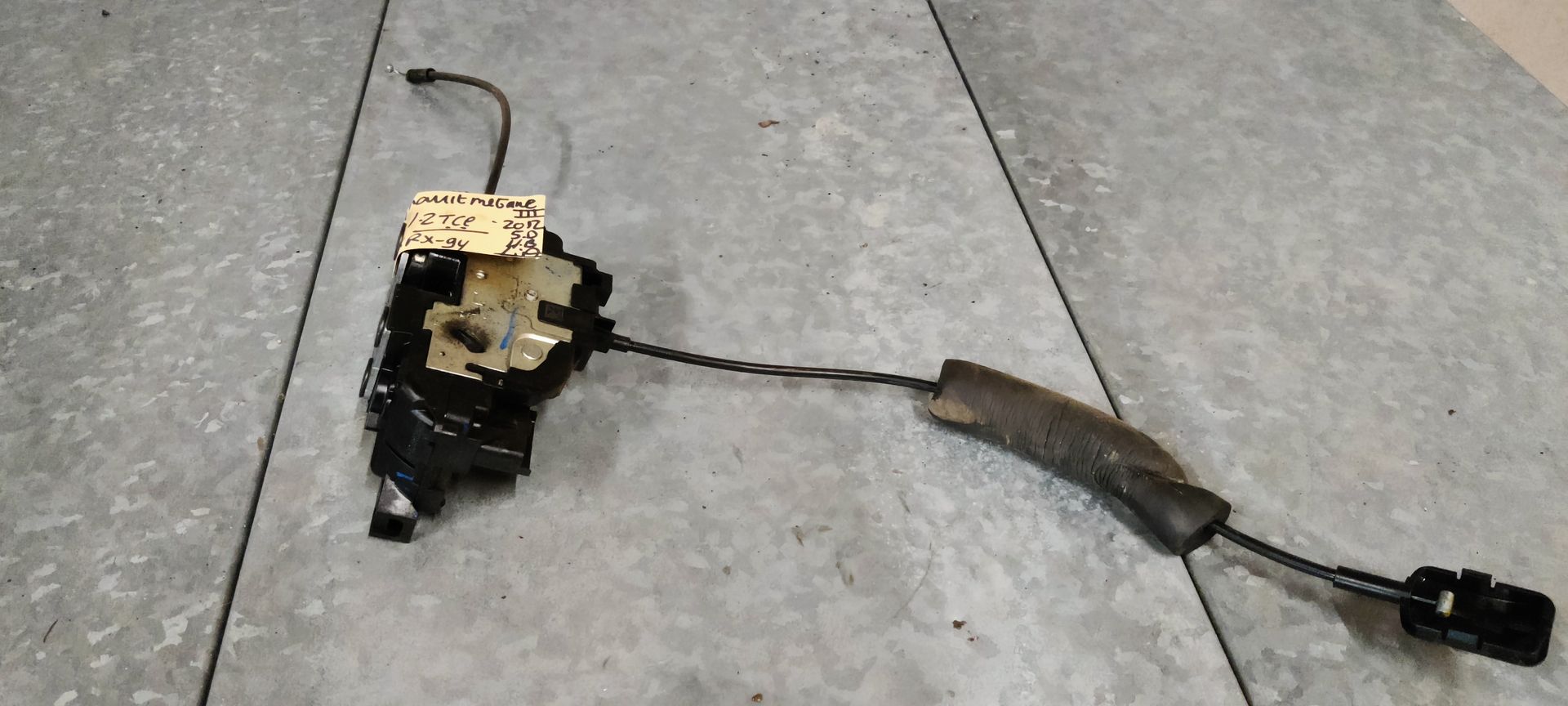 Een autodeurslotactuator met een aangesloten ontgrendelingskabel, rustend op een grijze tegelondergrond.