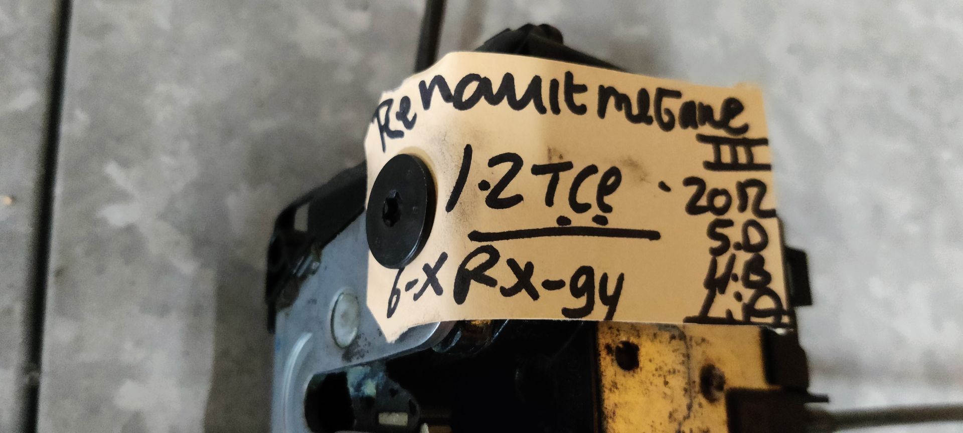 Een autodeurslotactuator met een aangesloten ontgrendelingskabel, rustend op een grijze tegelondergrond.