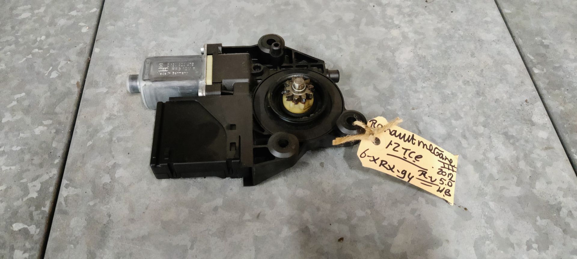 Een zwarte motor voor een elektrisch bedienbaar autoraam met tandwiel, geplaatst op een grijze ondergrond met een handgeschreven identificatielabel.