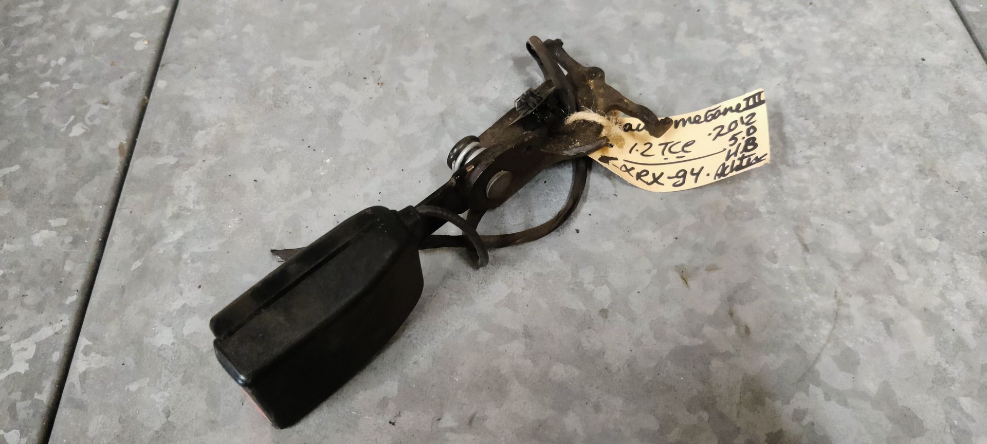 Een zwarte autogordelgesp met een metalen bevestigingsbeugel, rustend op een grijze tegelvloer.