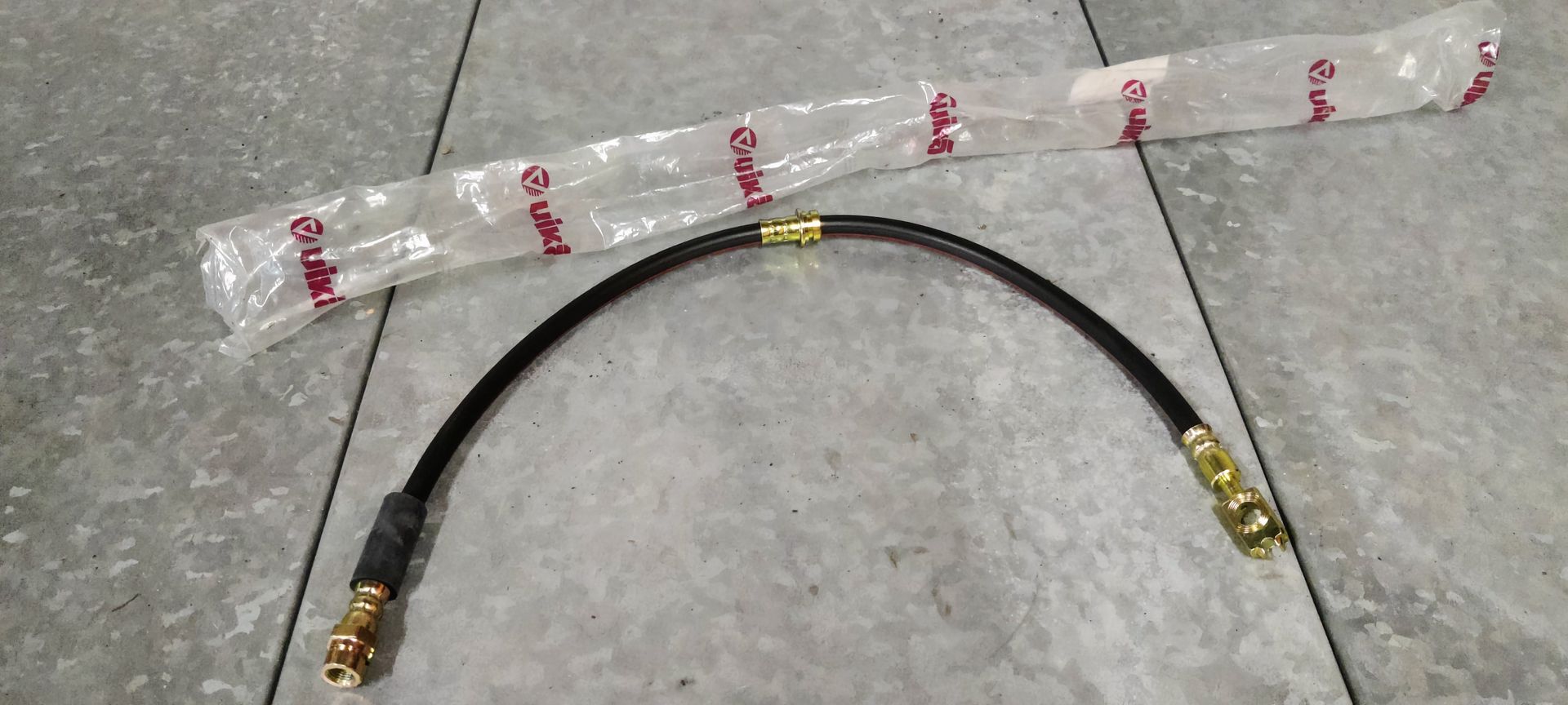 Een zwarte remslang voor auto's met messing schroefdraadaansluitingen, afgebeeld met de originele plastic verpakking.