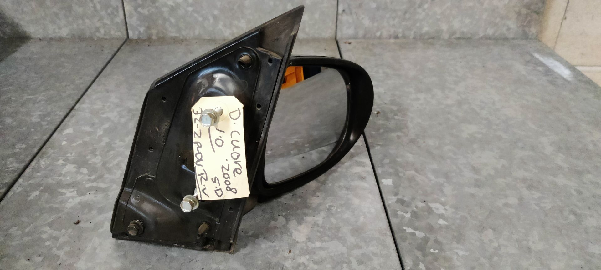 Een zwarte autospiegel met een aangehecht label, rustend op een metalen oppervlak.