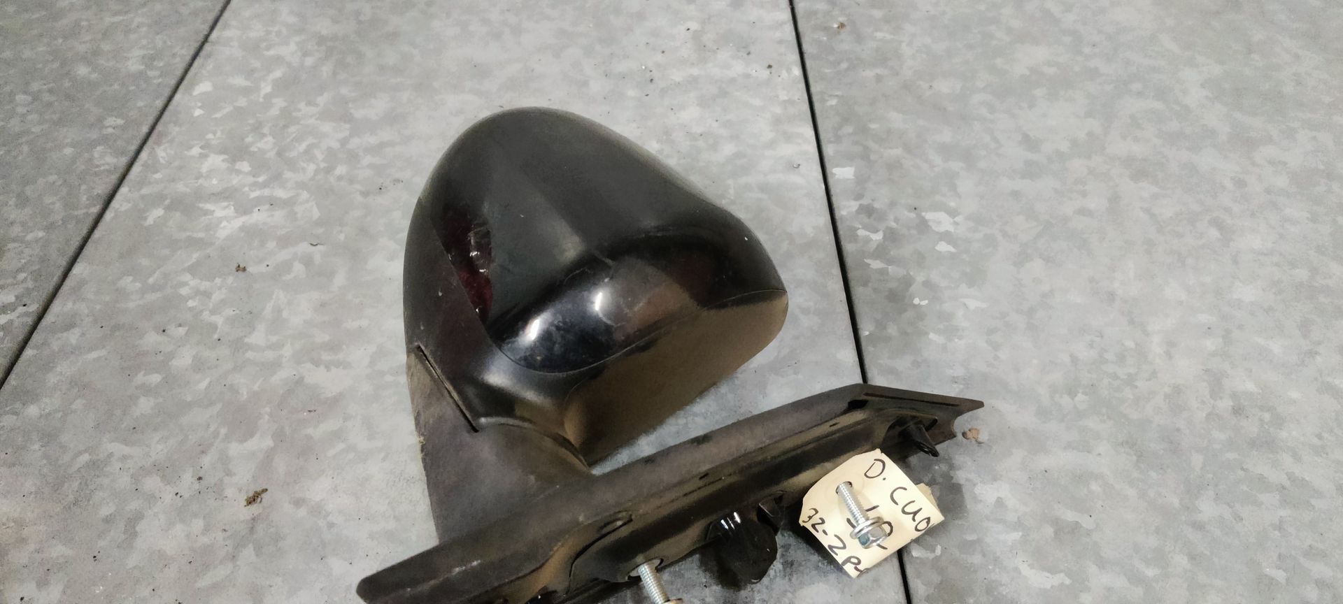 Een zwarte autospiegel met een aangehecht label, rustend op een metalen oppervlak.