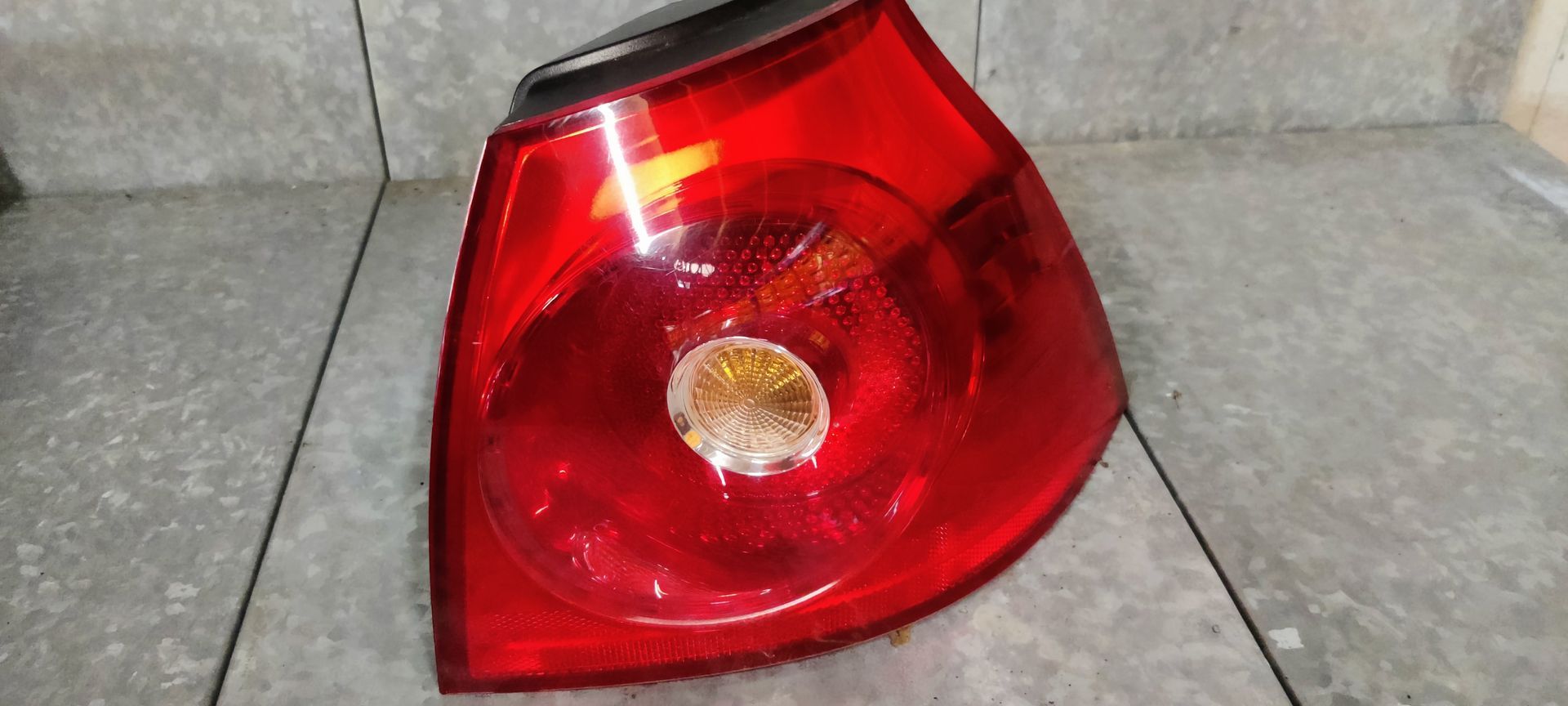 Een rode autoachterlichtunit met een ronde, witte lens voor het achteruitrijlicht in het midden, tegen een grijze achtergrond.