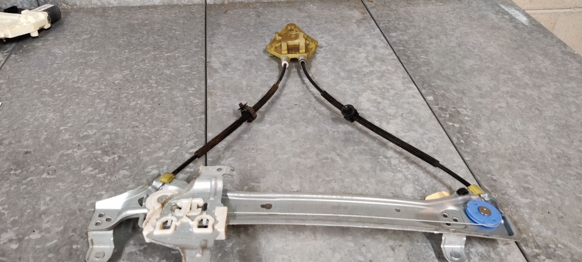 Een raammechanisme voor een auto, met metalen rails, zwarte kabels en een goudkleurige motorbehuizing, op een grijze tegelvloer.