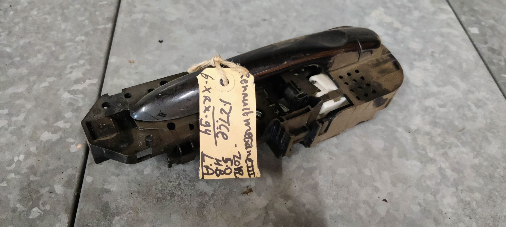Een zwarte buitendeurhendel van een auto met een geel identificatielabel, liggend op een grijze tegelvloer.