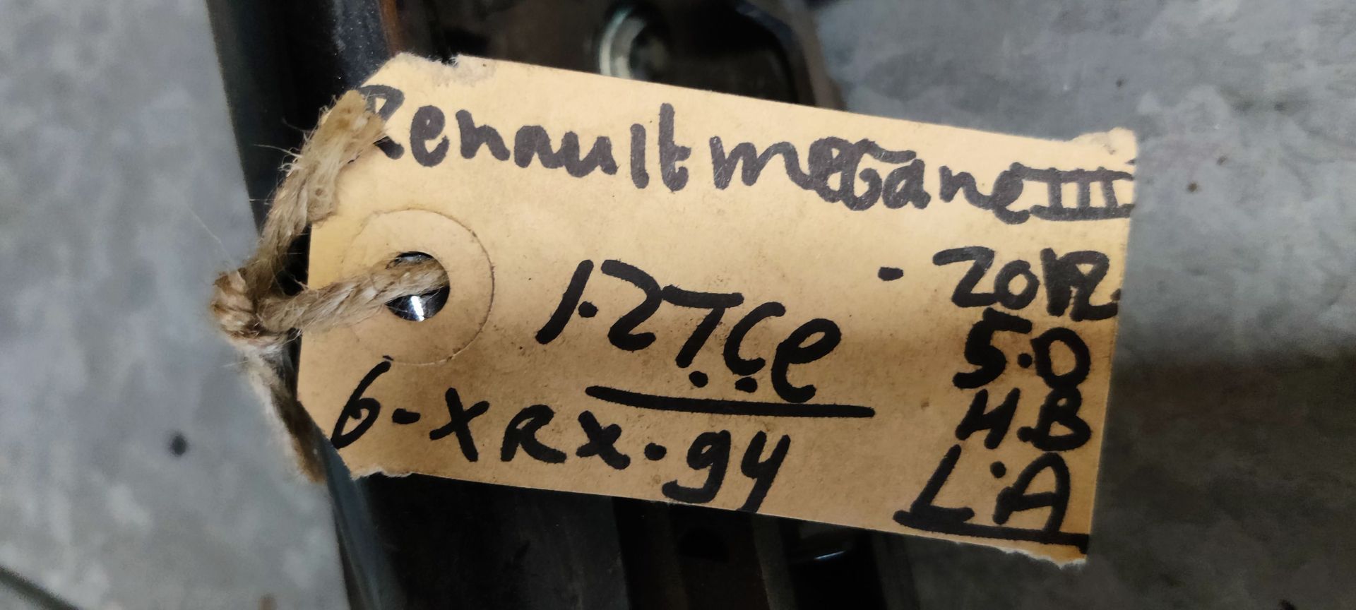 Een zwarte buitendeurhendel van een auto met een geel identificatielabel, liggend op een grijze tegelvloer.