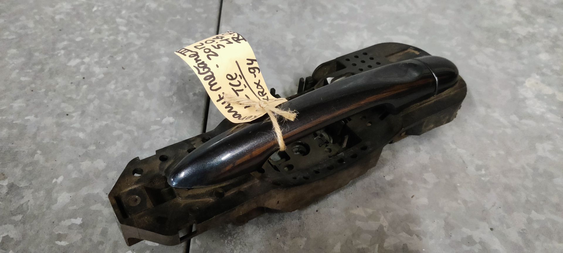 Een donkere autodeurhendel met een handgeschreven papieren label eraan, liggend op een grijze betonnen ondergrond.