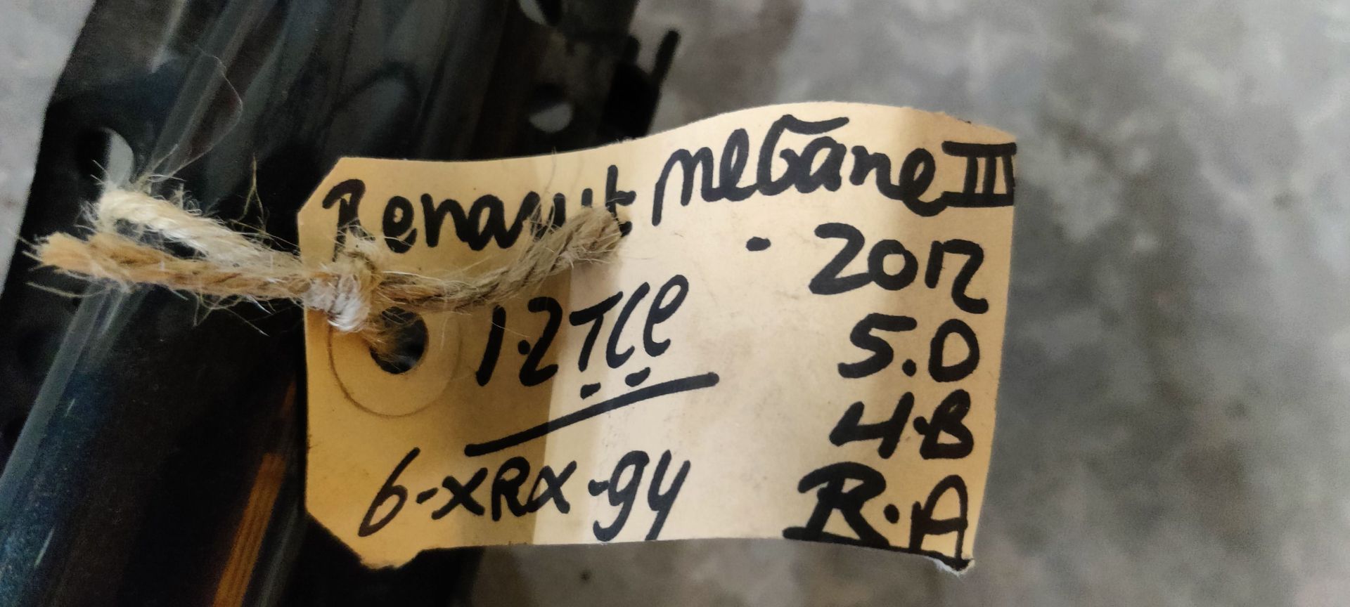 Een donkere autodeurhendel met een handgeschreven papieren label eraan, liggend op een grijze betonnen ondergrond.