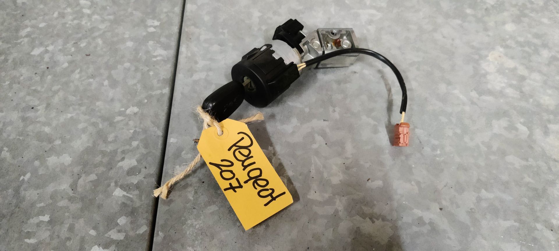 Een zwart contactslot en sleutel van een Peugeot 207 met een geel identificatielabel op een grijze ondergrond.