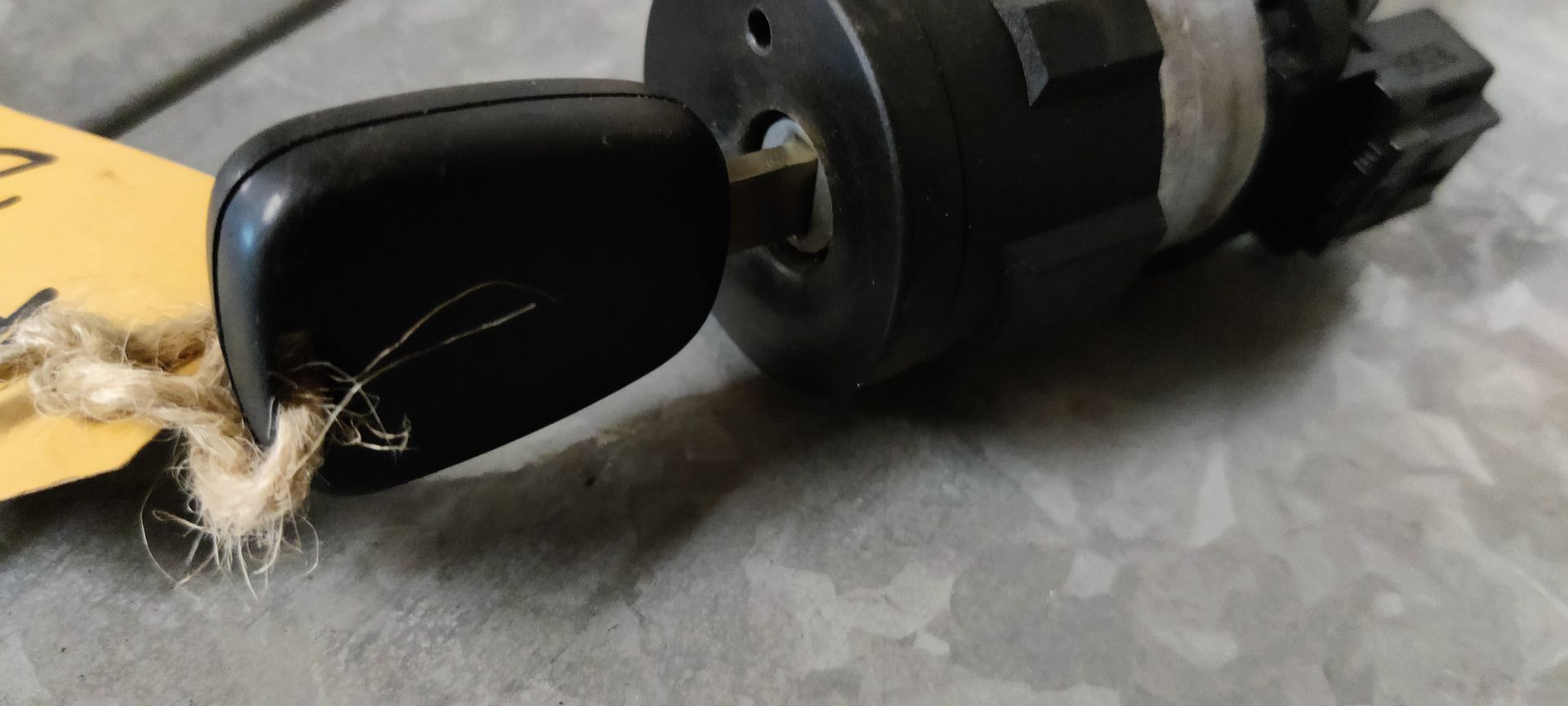 Een zwart contactslot en sleutel van een Peugeot 207 met een geel identificatielabel op een grijze ondergrond.