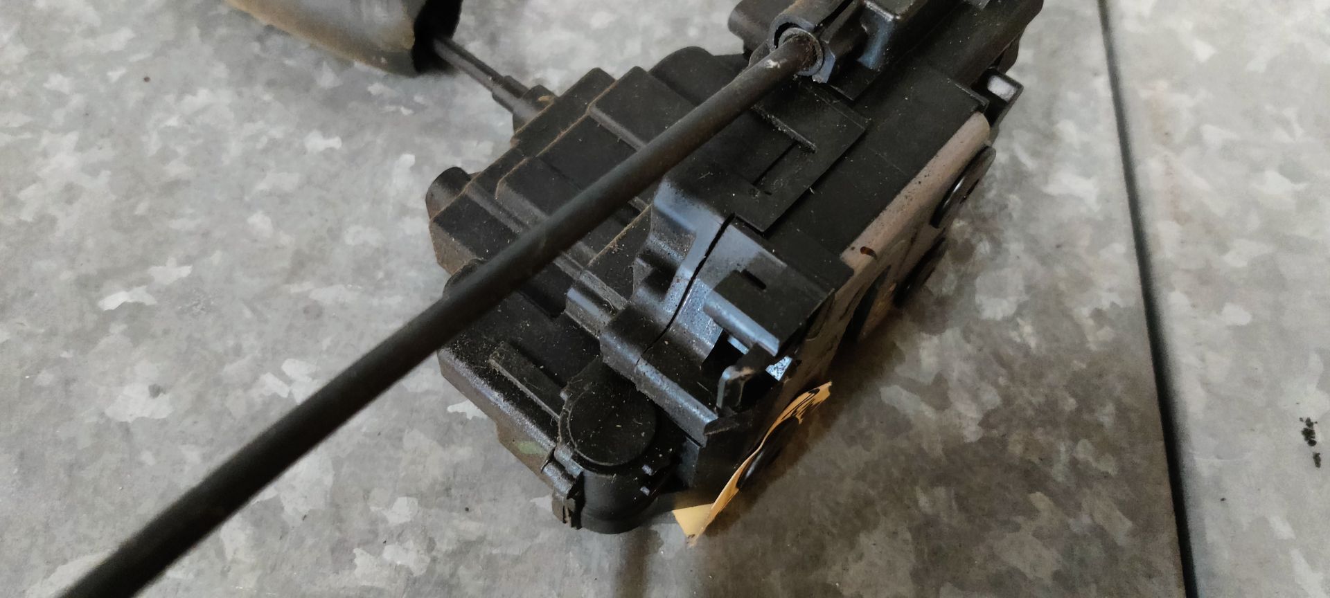 Een actuatormechanisme voor een autodeurslot met aangesloten kabels, afgebeeld tegen een grijze, gestructureerde achtergrond.