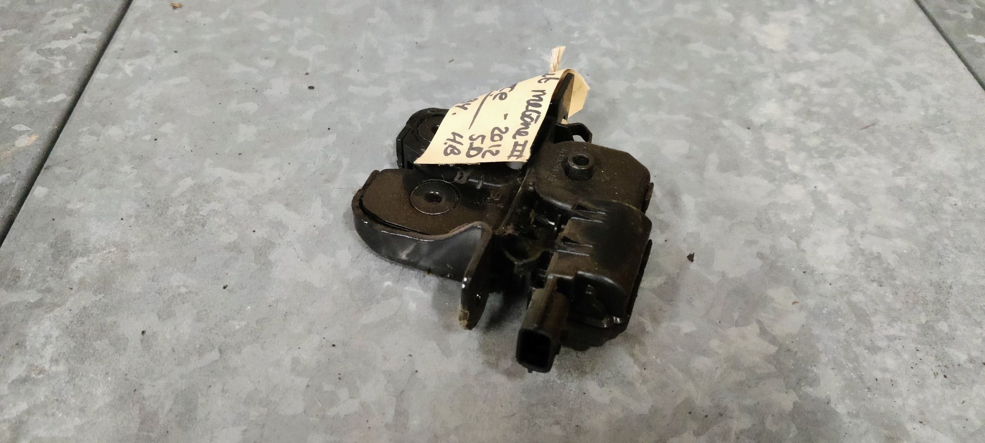 Een zwart metalen autodeurslot met een aangehecht identificatielabel, rustend op een grijze ondergrond.