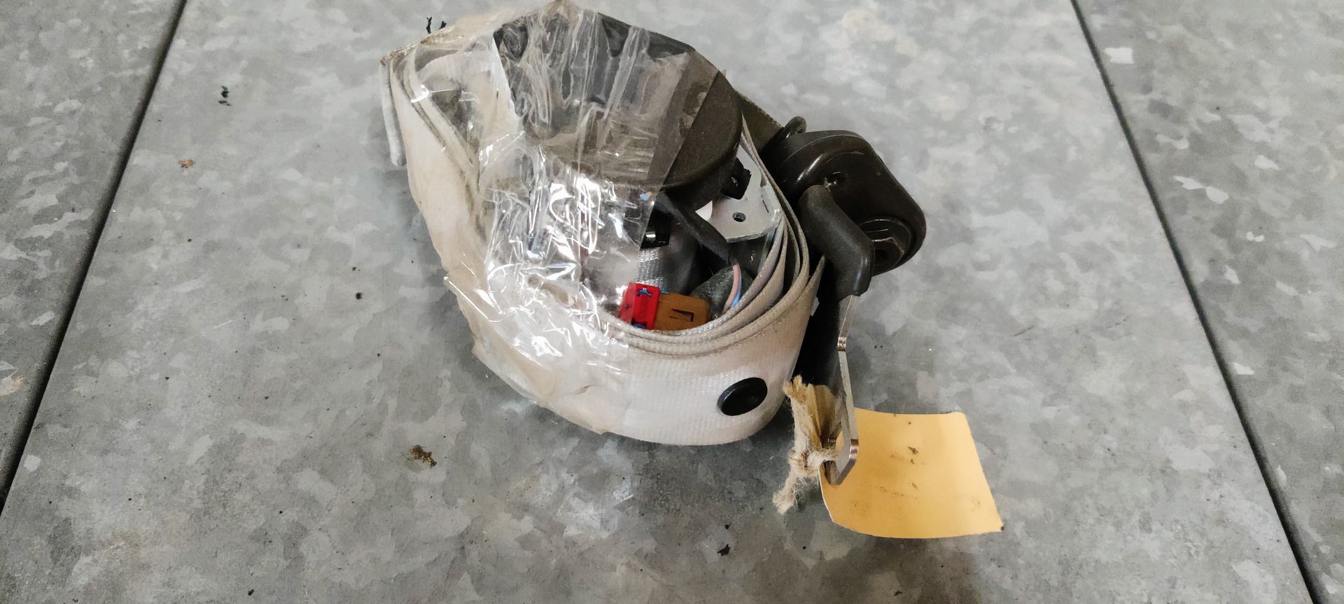 Een wit oprolmechanisme voor autogordels met een geel label, rustend op een grijze betonnen ondergrond.