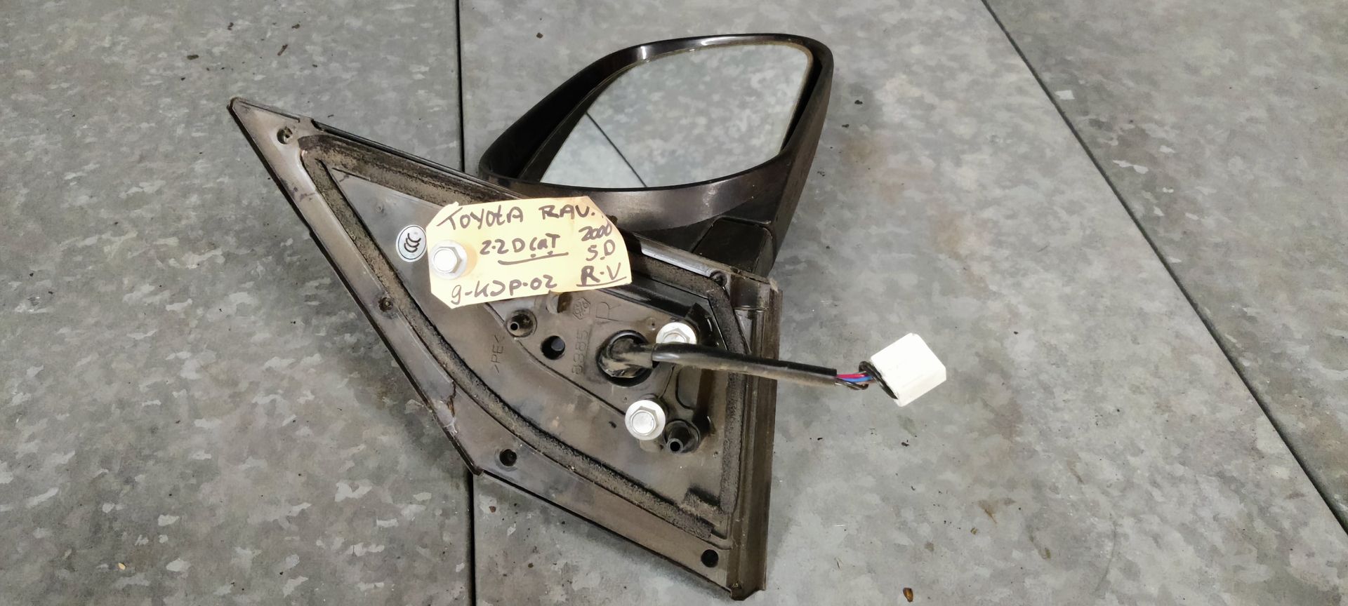 Een autospiegel met aangesloten kabelboom, rustend op een betonnen ondergrond met een handgeschreven label eraan.