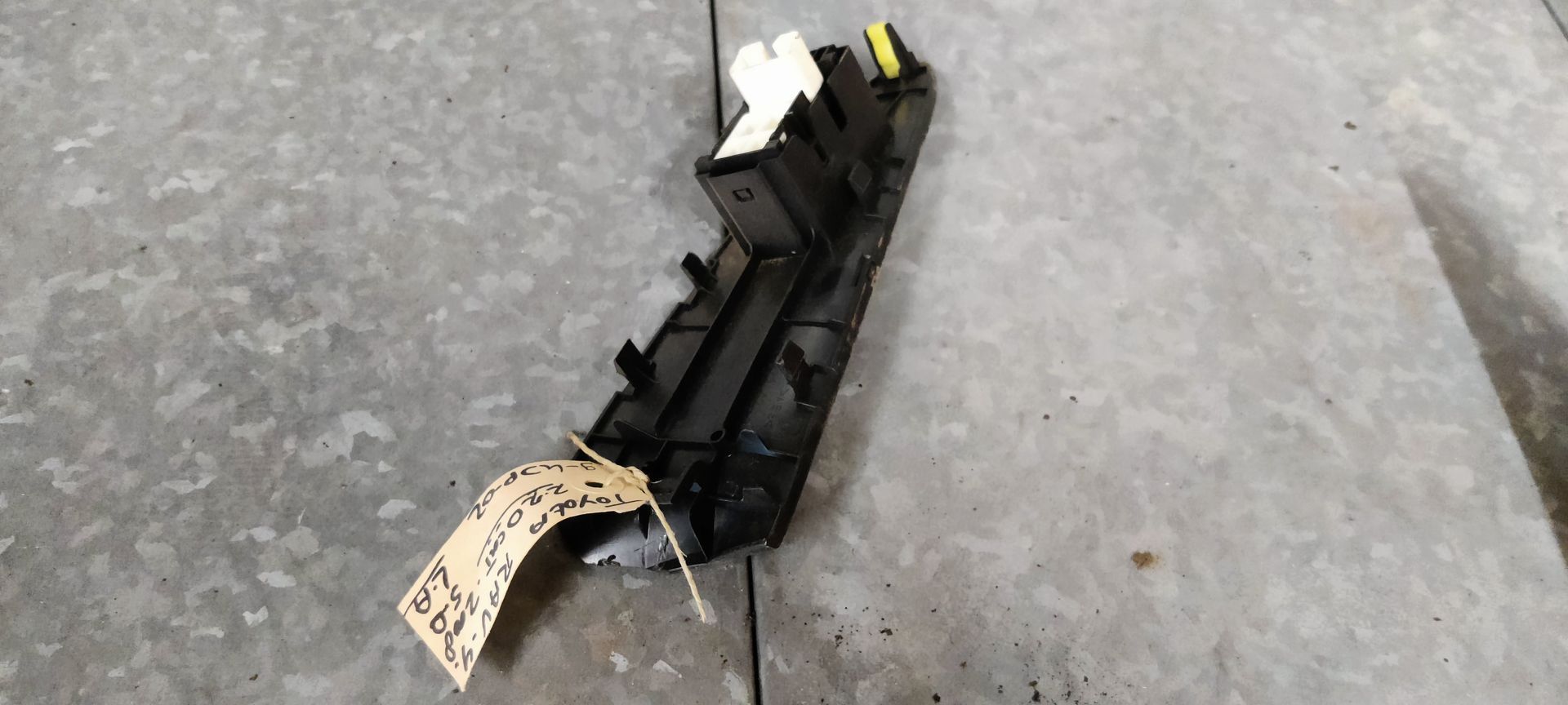 Een zwarte autodeurhendel met een aangehecht identificatielabel, rustend op een grijze betonnen ondergrond.