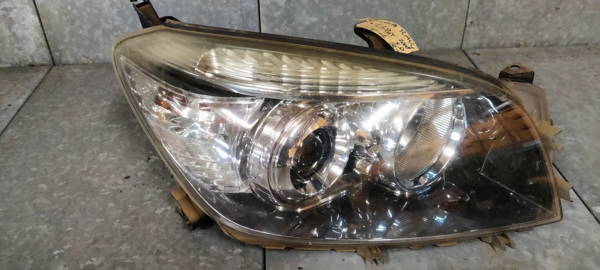 Een gebruikte autokoplamp met een heldere lens en een ronde projectorlamp, geplaatst tegen een grijze tegelachtergrond.