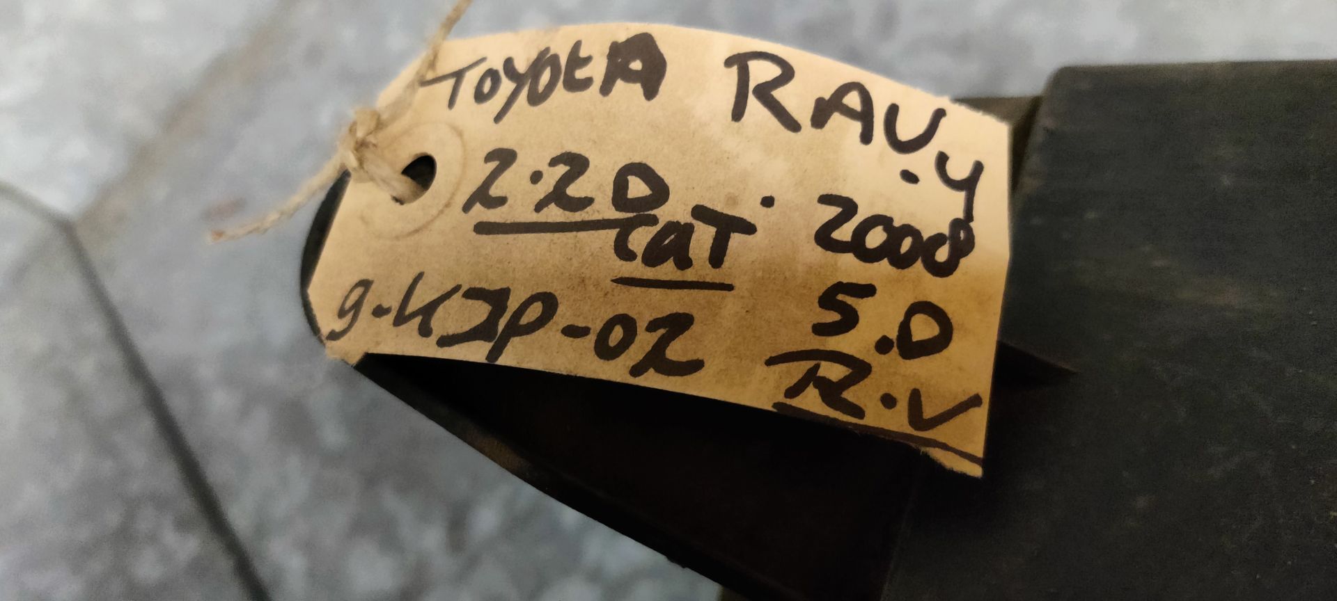 Een gebruikte autokoplamp met een heldere lens en een ronde projectorlamp, geplaatst tegen een grijze tegelachtergrond.