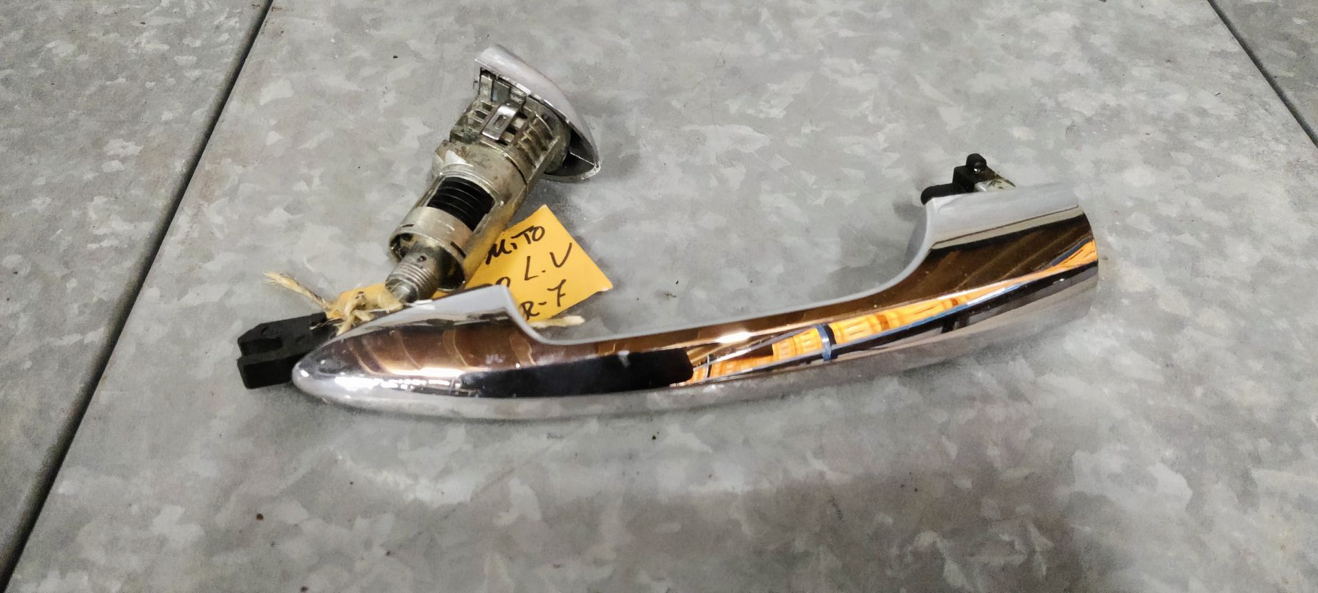 Alfa Romeo MiTo deurklink links. Een verchroomde autodeurhendel met een ingebouwd slotcilindermechanisme, rustend op een grijze tegelondergrond.