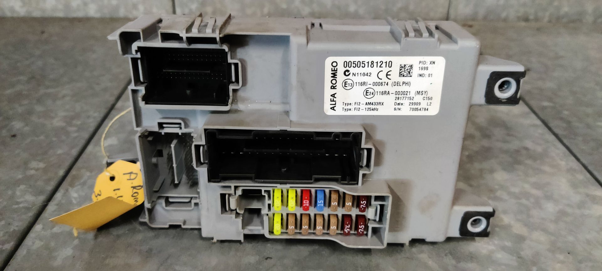 Een grijze zekeringkast en carrosserieregelmodule voor auto's met twee grote zwarte connectorpoorten en een rij gekleurde zekeringen.