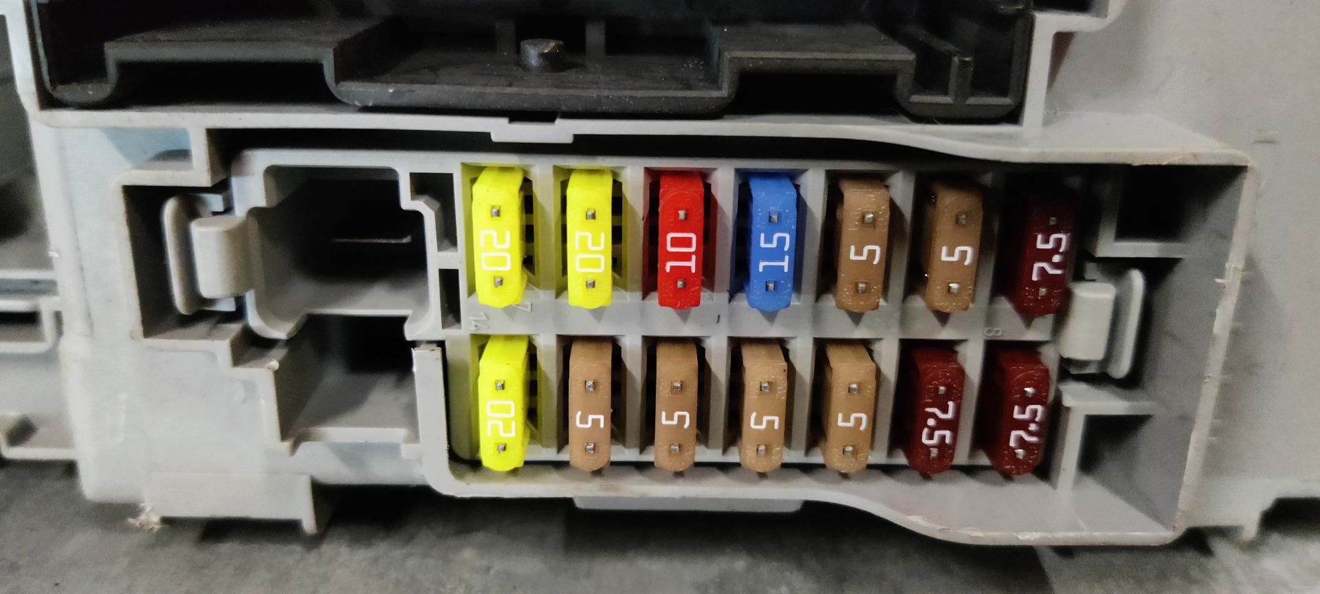 Een grijze zekeringkast en carrosserieregelmodule voor auto's met twee grote zwarte connectorpoorten en een rij gekleurde zekeringen.