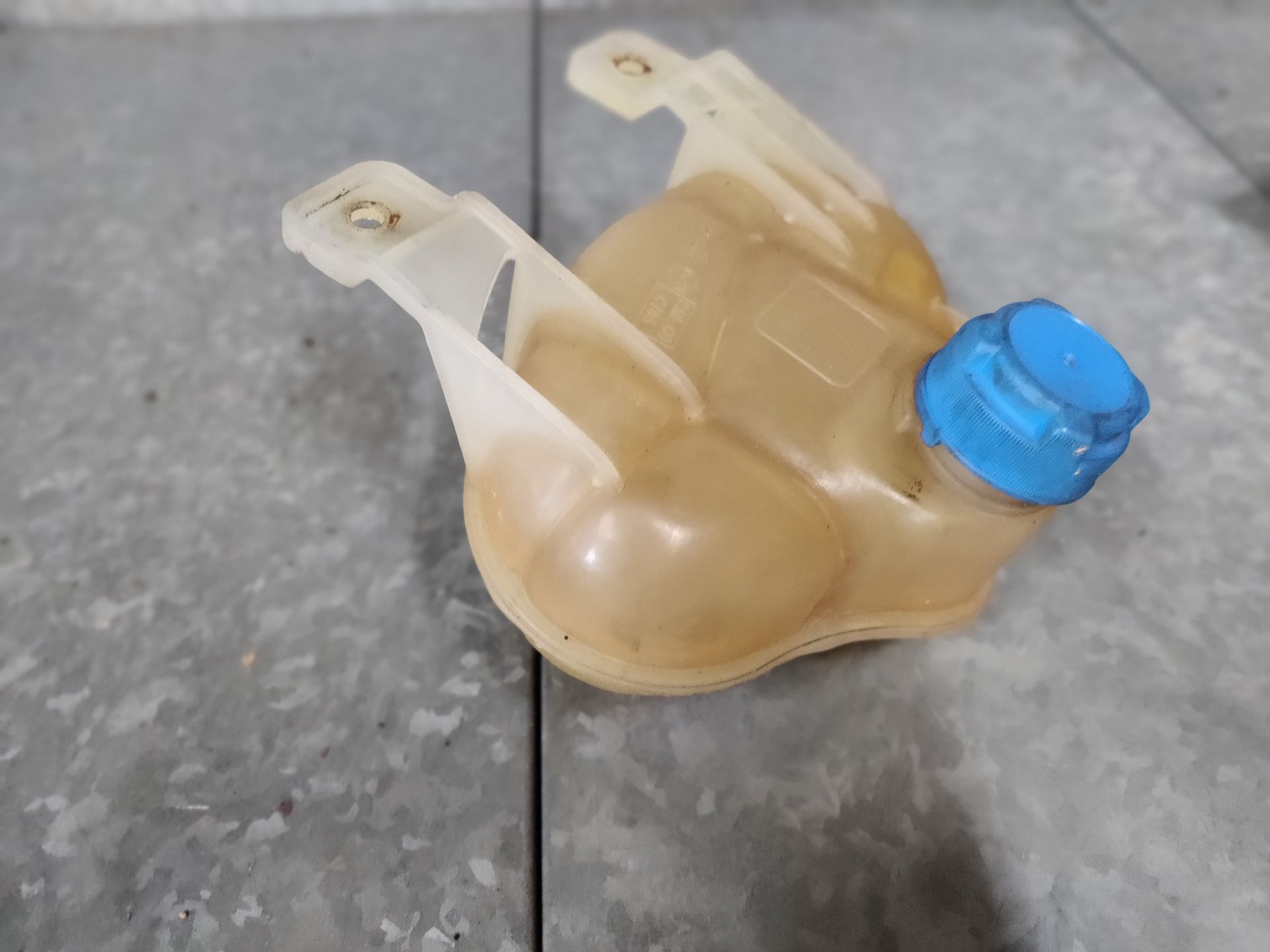 Een vergeeld, doorschijnend plastic expansievat voor autokoelvloeistof met een blauwe dop, rustend op een grijze tegelvloer.