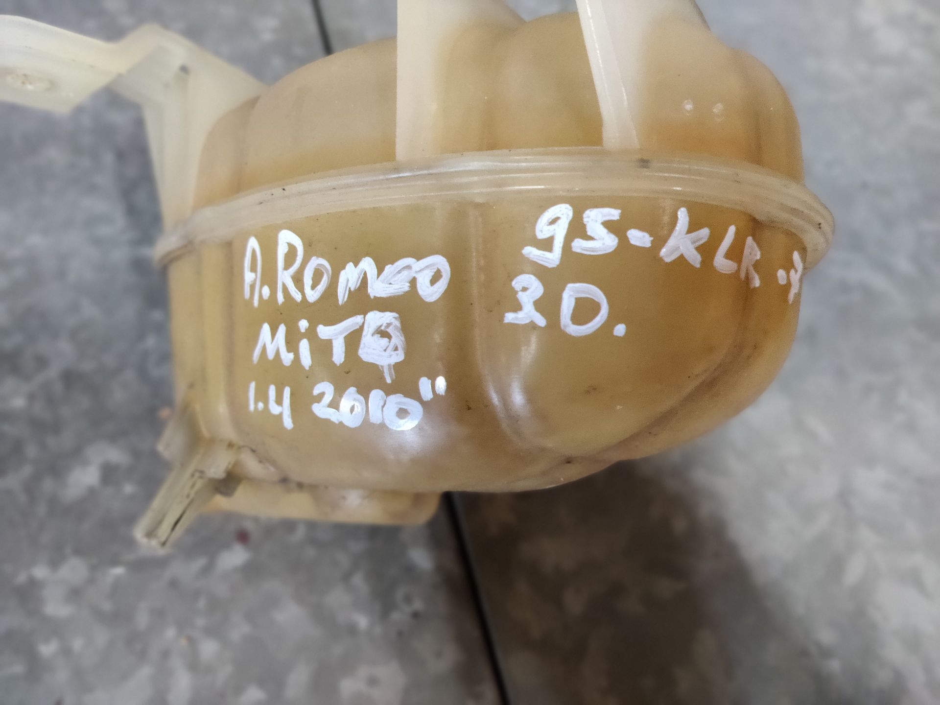 Een vergeeld, doorschijnend plastic expansievat voor autokoelvloeistof met een blauwe dop, rustend op een grijze tegelvloer.