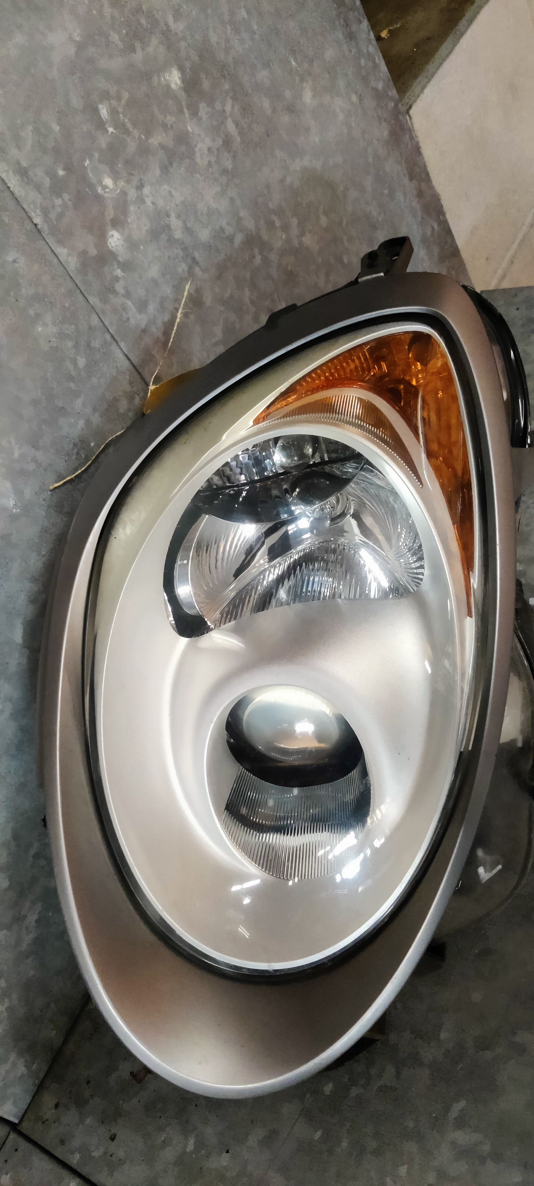 Een gedemonteerde koplampunit van een auto met projectorlens, reflector en amberkleurig knipperlichtgedeelte op een grijze vloer.