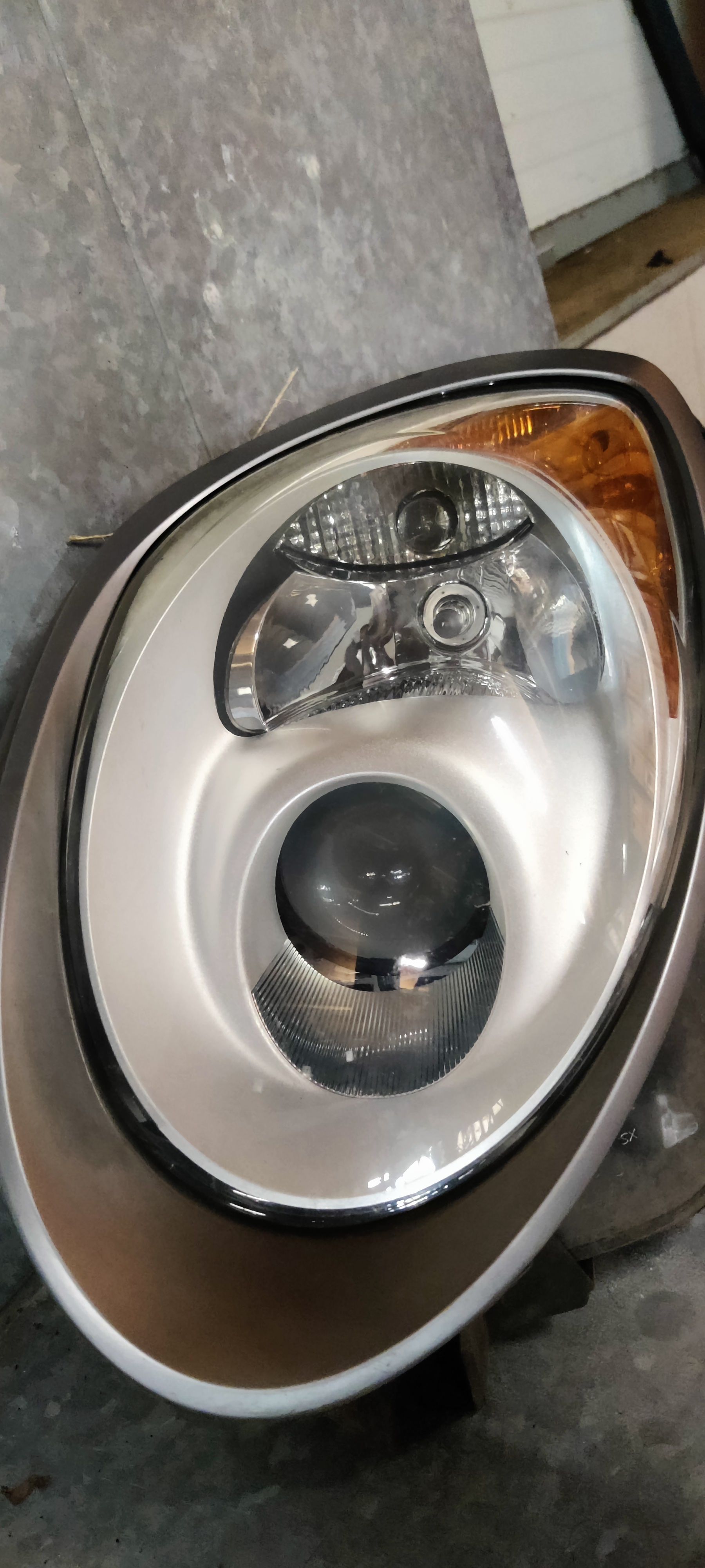 Een gedemonteerde koplampunit van een auto met projectorlens, reflector en amberkleurig knipperlichtgedeelte op een grijze vloer.