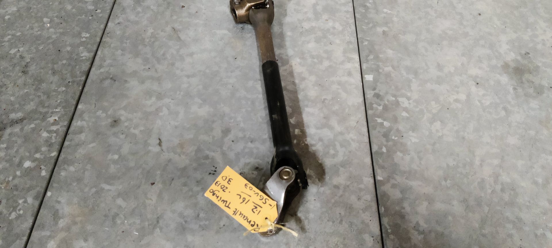 Een stuurkolom van een auto met kruiskoppelingen aan beide uiteinden, liggend op een grijze betonnen vloer met een identificatielabel.