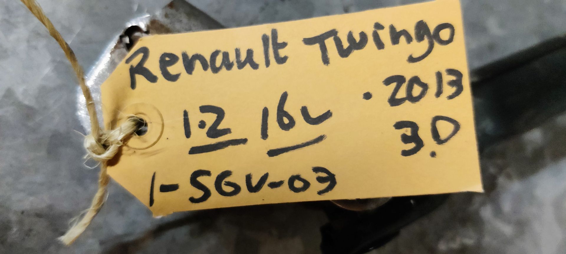 Een stuurkolom van een auto met kruiskoppelingen aan beide uiteinden, liggend op een grijze betonnen vloer met een identificatielabel.