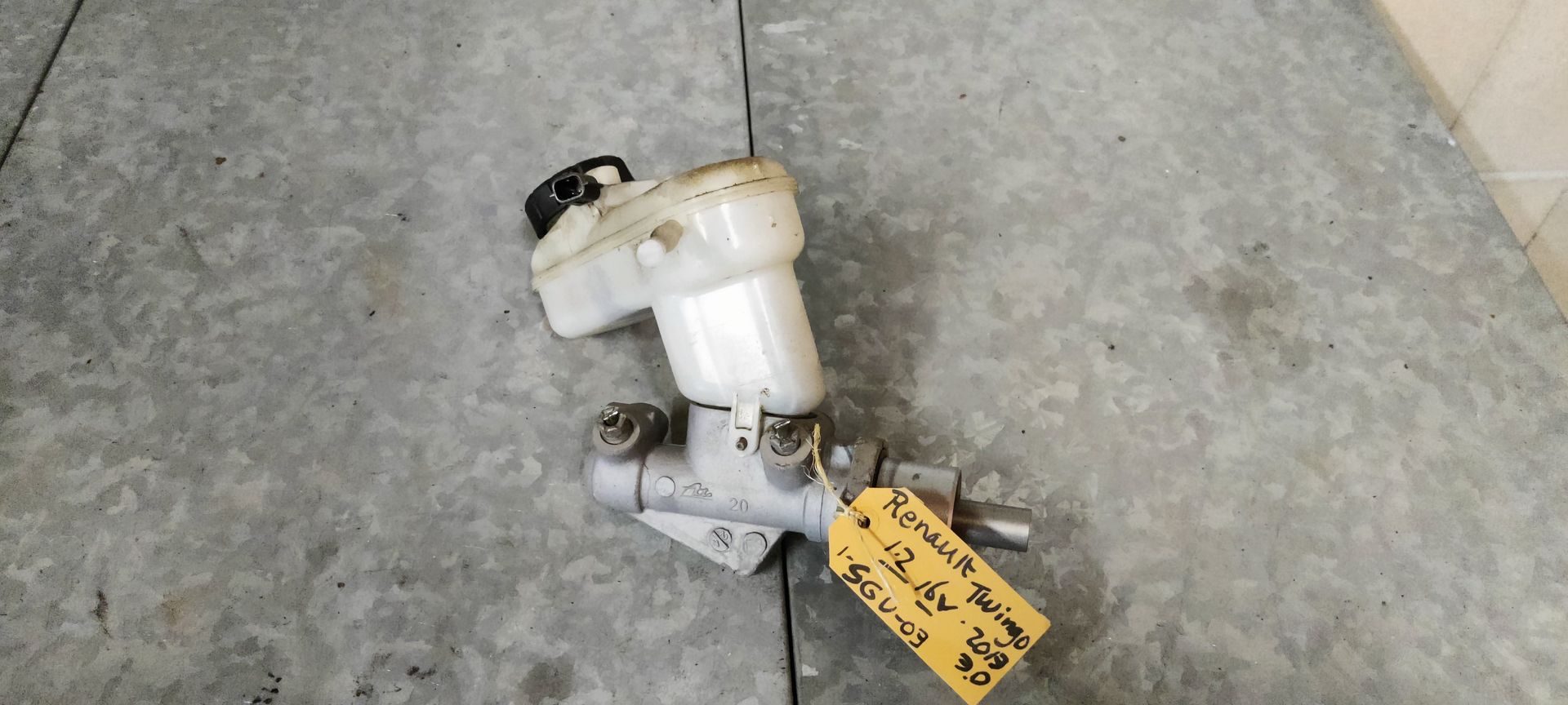 Een gebruikte hoofdremcilinder van een auto met een wit plastic vloeistofreservoir, staand op een grijze betonnen ondergrond.