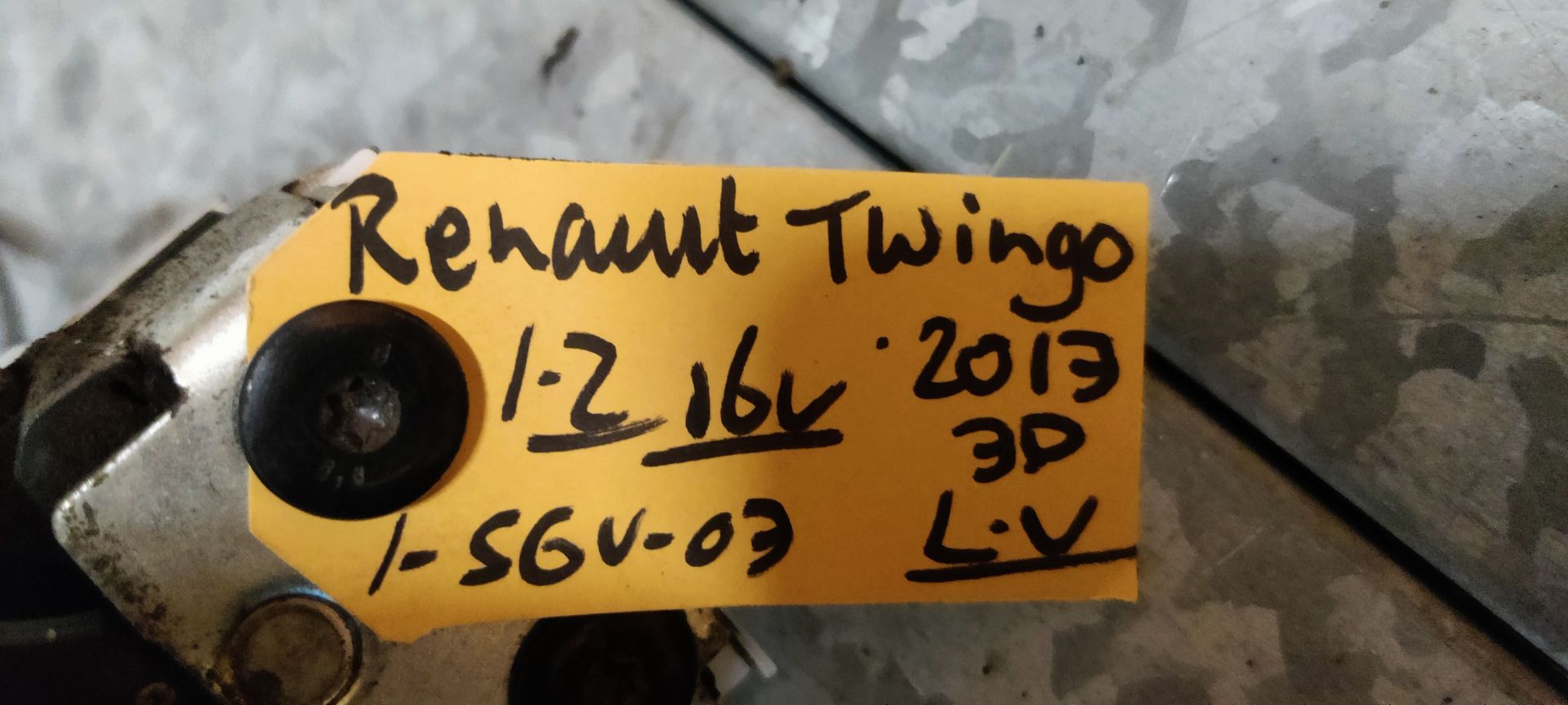 Een metalen deurslotmechanisme voor een Renault Twingo, met een geel identificatielabel op een grijs oppervlak.