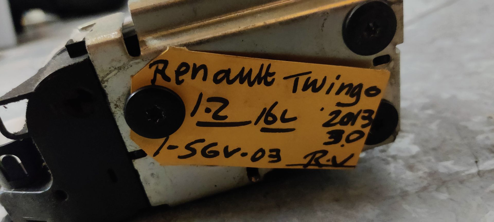 Een deurslotmechanisme van een Renault-voertuig met een geel identificatielabel, rustend op een grijze tegelondergrond.