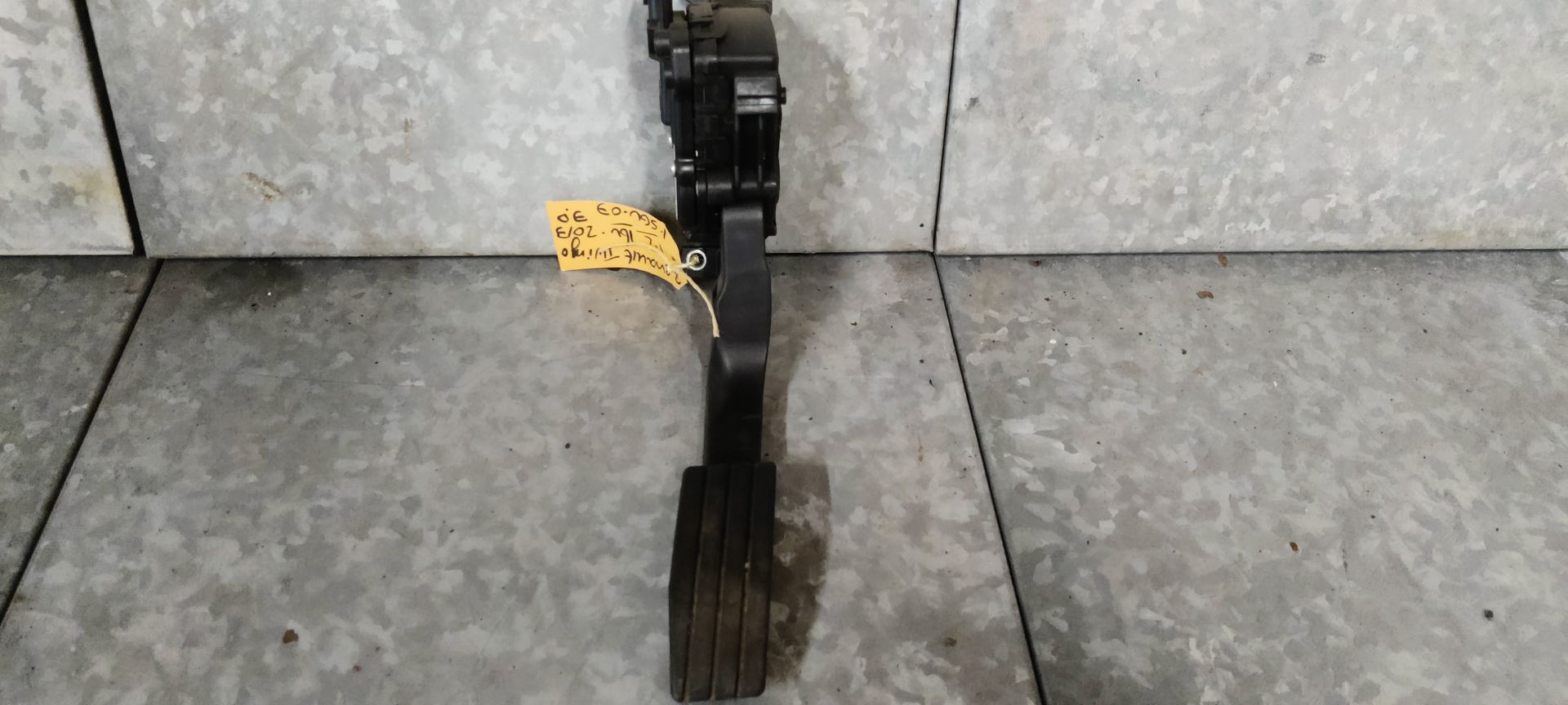 Een zwart elektronisch gaspedaal voor een voertuig met een geel identificatielabel op een grijs oppervlak.