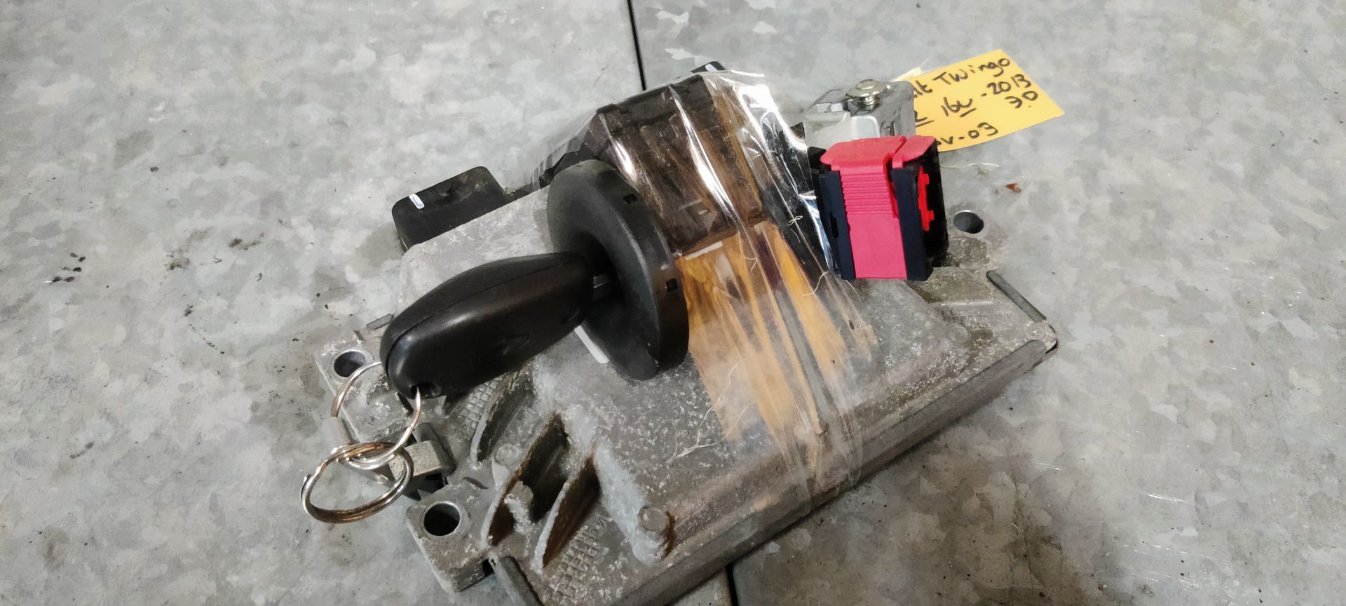 Een metalen ECU-module voor auto's met een zwarte sleutel die in een poort is gestoken, een rode connector en een geel label.
