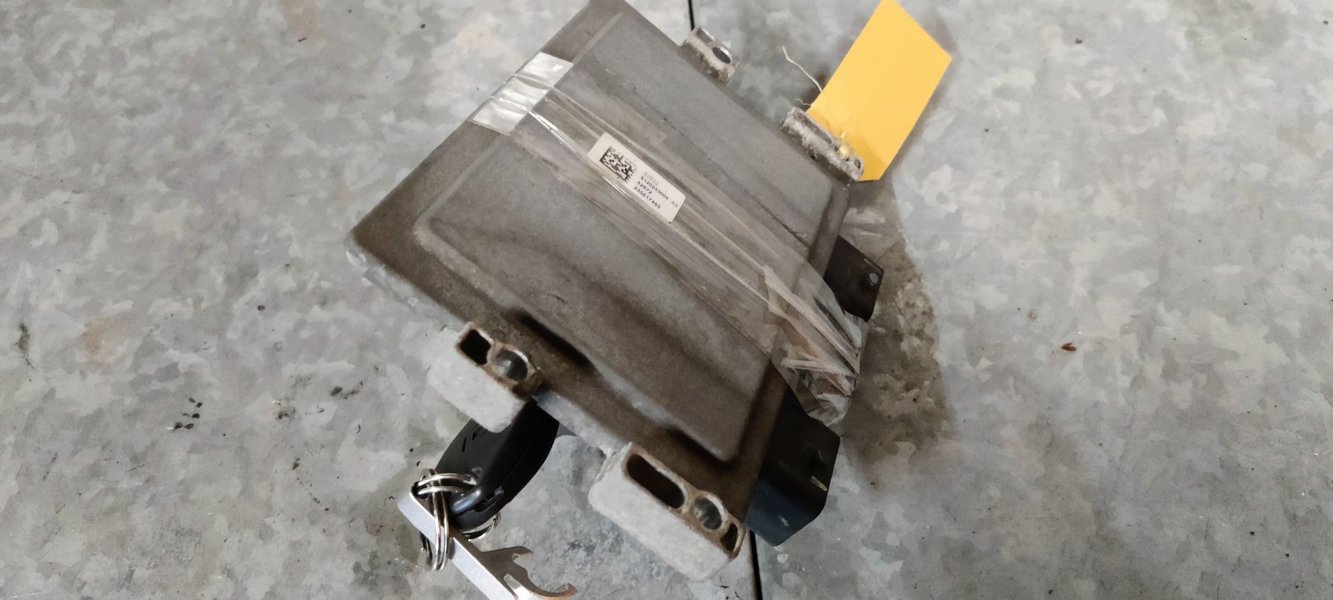 Een metalen ECU-module voor auto's met een zwarte sleutel die in een poort is gestoken, een rode connector en een geel label.