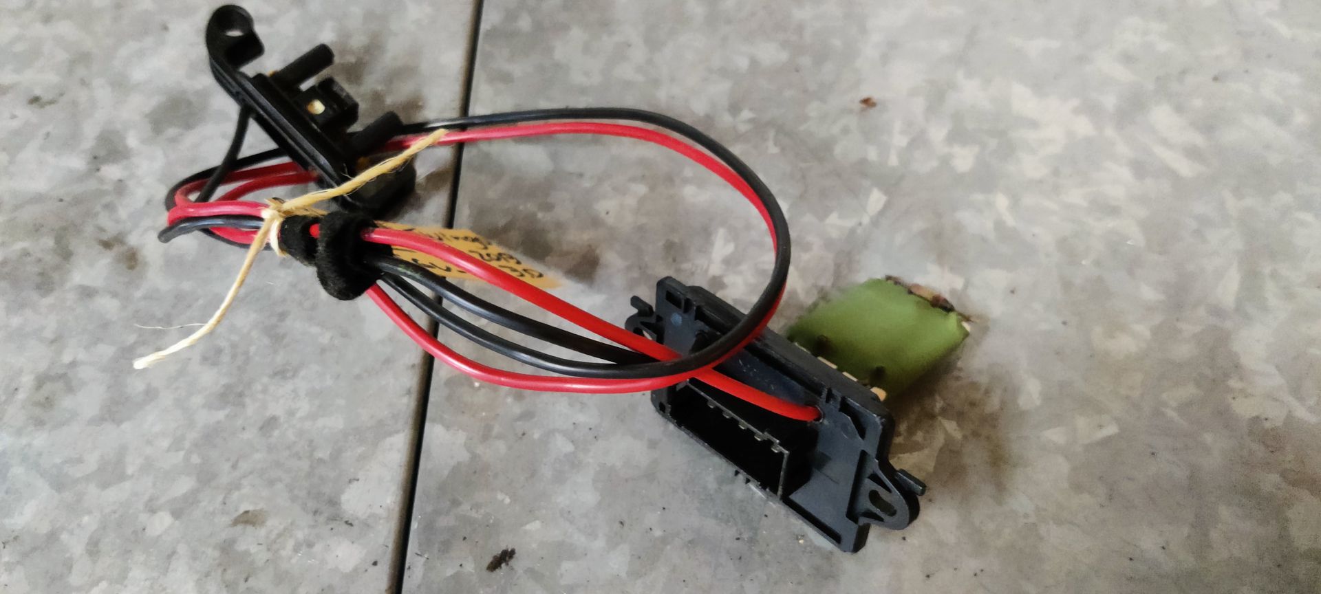 Een weerstand van een autoventilator met kabelboom en plastic connector, rustend op een grijze tegelondergrond.