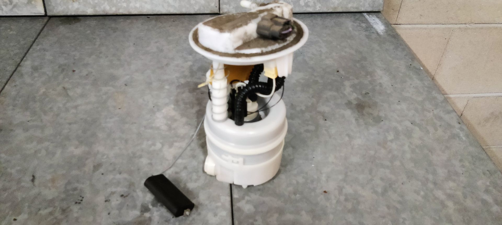 Een gebruikte brandstofpompunit voor auto's met een losse rechthoekige sensor, staand op een grijze betonnen ondergrond.