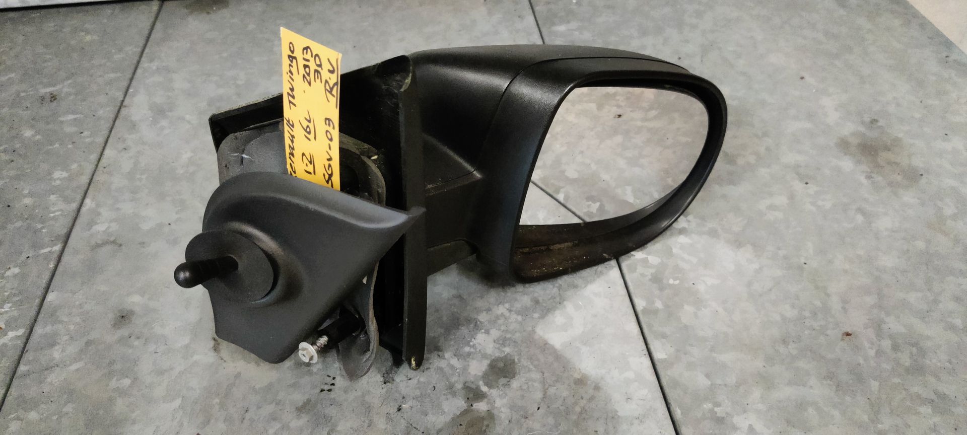 Een zwarte, handmatig verstelbare autospiegel met een geel identificatielabel, rustend op een grijze betonnen vloer.