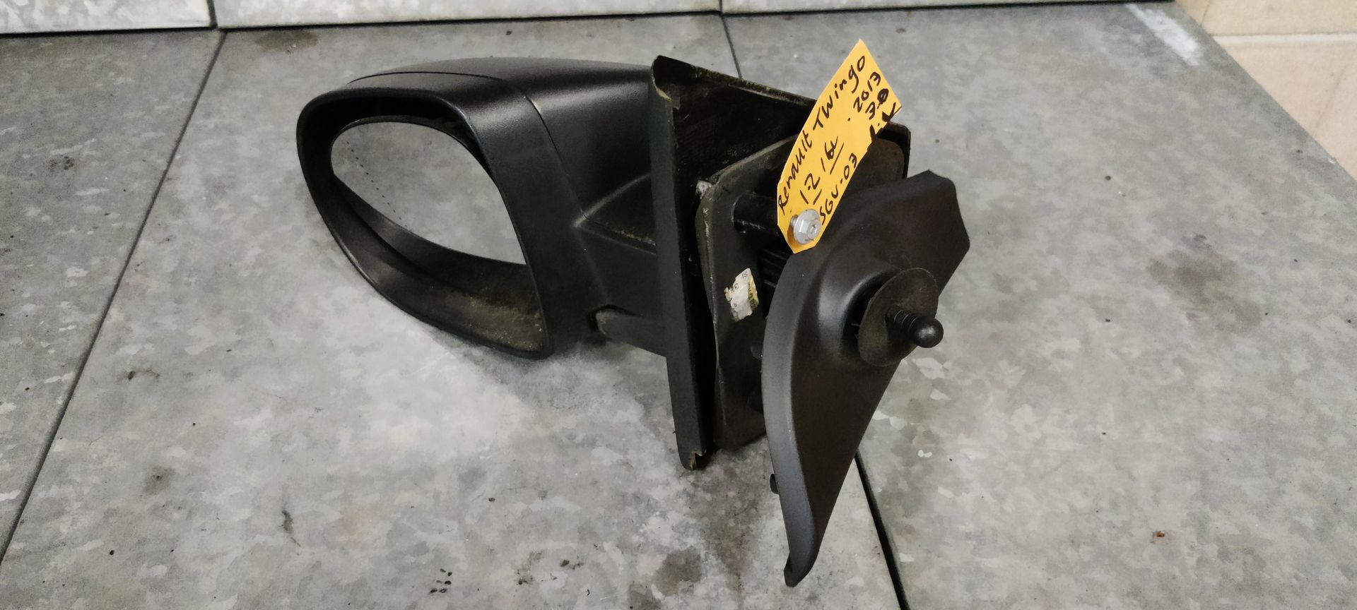 Zwarte zijspiegel van een auto met een geel identificatielabel, rustend op een grijze betonnen vloer.