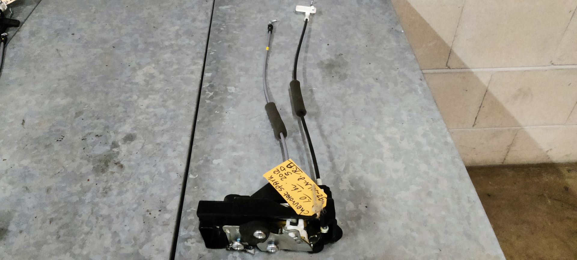 Een autodeurvergrendeling met aangesloten trekdraden en een geel identificatielabel, rustend op een grijze werkbank.