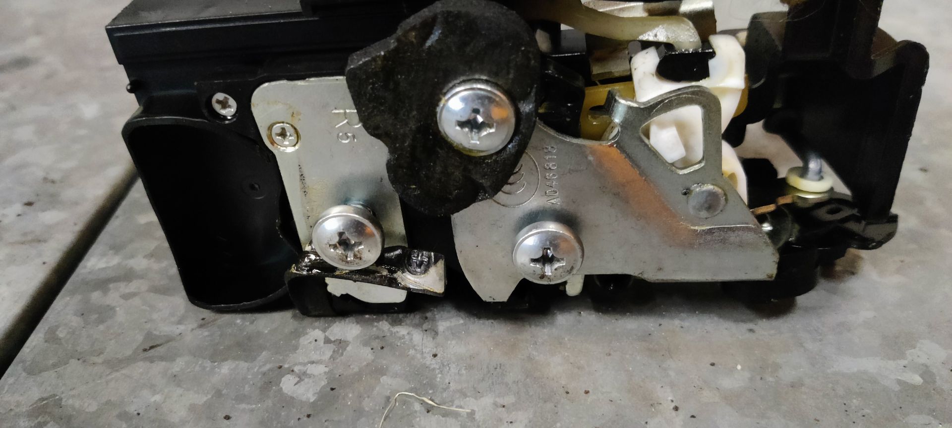 Een autodeurvergrendeling met aangesloten trekdraden en een geel identificatielabel, rustend op een grijze werkbank.