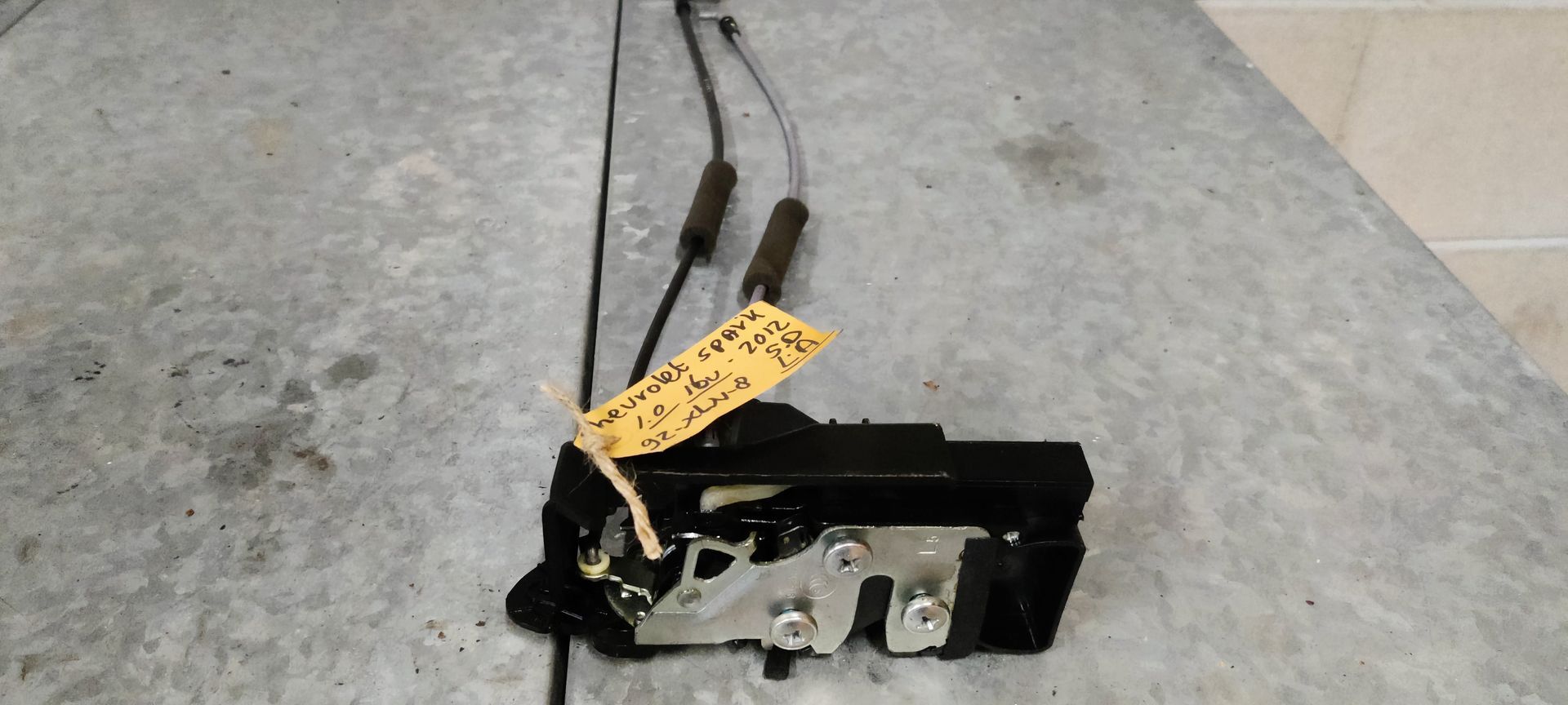 Een autodeurvergrendeling met twee aangesloten kabels, geplaatst op een grijs metalen oppervlak.