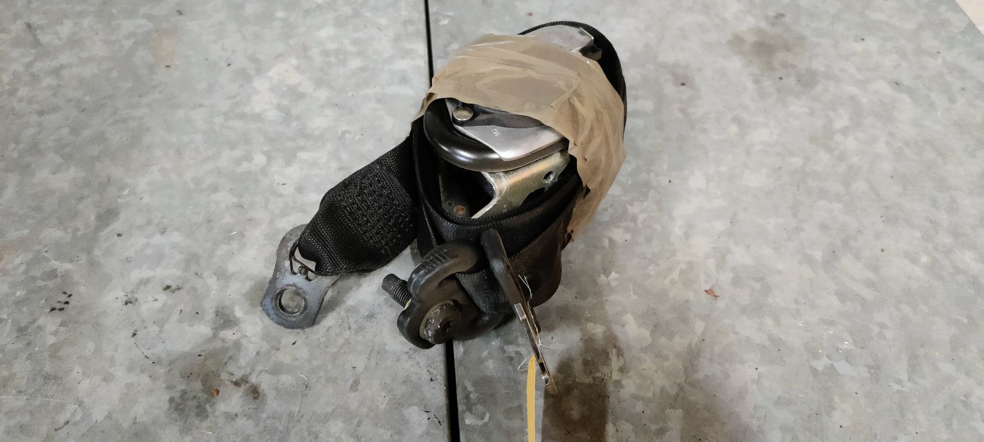 Een metalen oprolmechanisme voor autogordels met zwarte band en beige tape, rustend op een grijze betonnen vloer.
