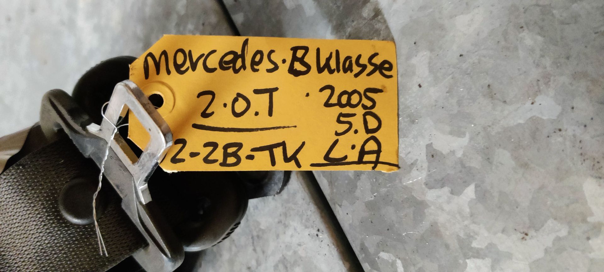 Een metalen oprolmechanisme voor autogordels met zwarte band en beige tape, rustend op een grijze betonnen vloer.