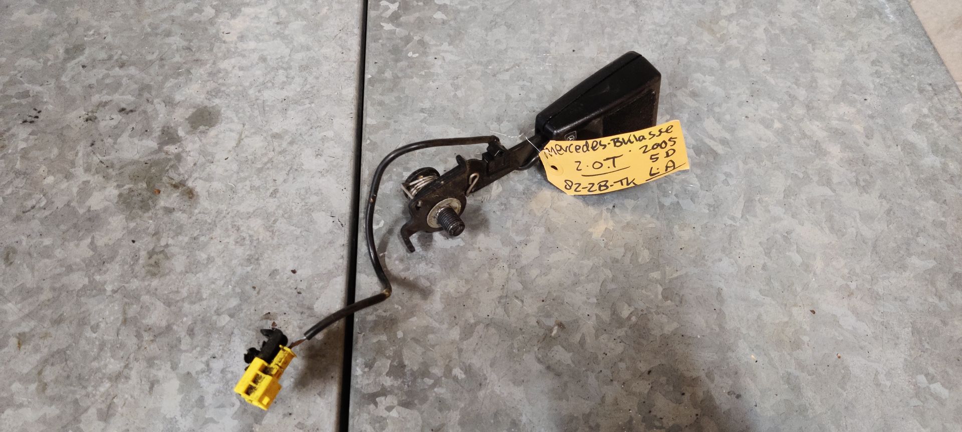 Een zwarte autogordelgesp met bedrading en een gele connector, liggend op een grijze betonnen vloer met een geel label.