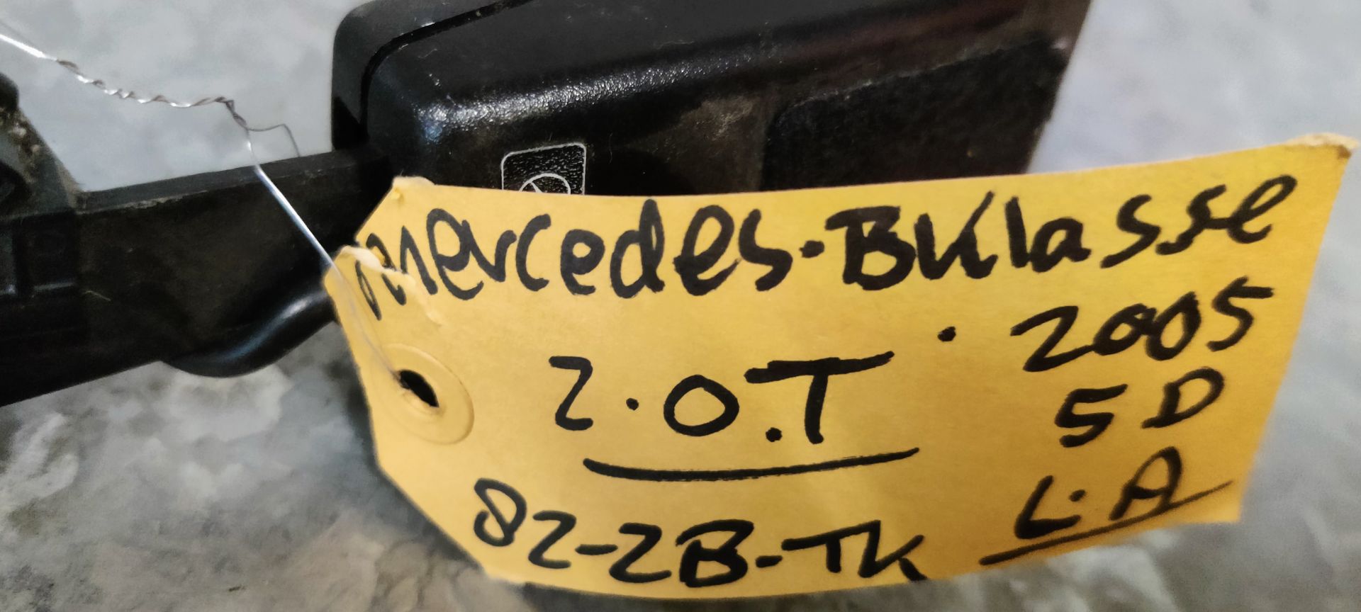 Een zwarte autogordelgesp met bedrading en een gele connector, liggend op een grijze betonnen vloer met een geel label.