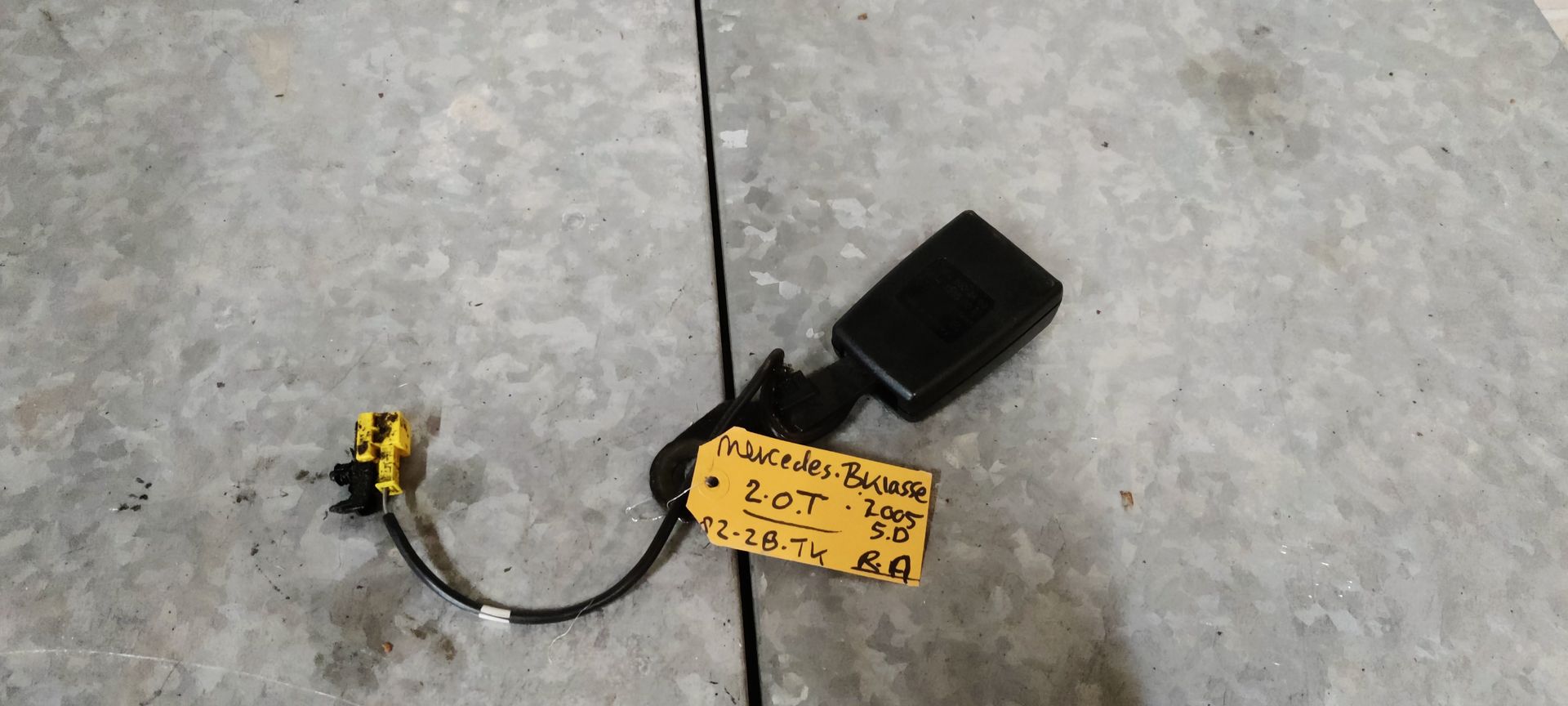 Een zwarte autogordelgesp met een gele elektrische connector en een handgeschreven identificatielabel.